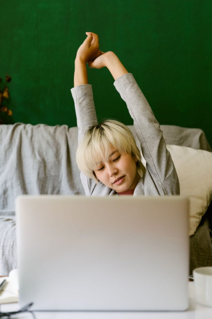 woman working at a laptop in bed and stretching her arms above her head in front of a green wall